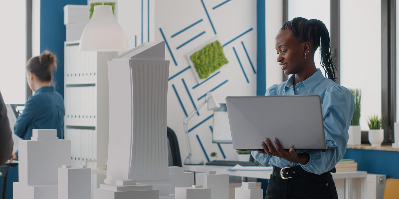 Woman architect holding laptop to analyze building model in architectural office. Engineer working with computer and maquette to design construction structure for urban project.